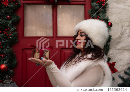 Christmas gift woman holding small present by festive red door in white winter outfit Christmas gift woman holding small present by festive red door in white winter outfit 135362614