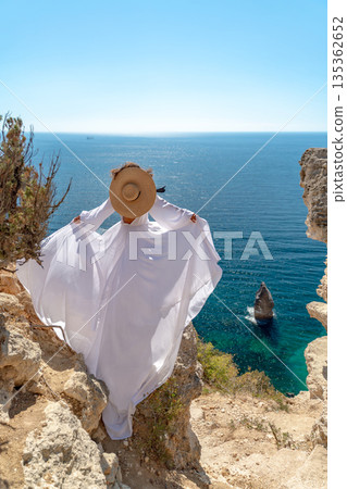 Woman, sea, cliff. Woman in flowing white dress, straw hat, stands on cliff overlooking vast blue sea. Summer travel freedom. 135362652