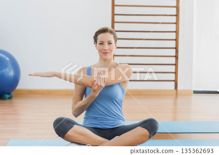 Woman stretching right arm across chest while holding left wrist on blue mat in exercise studio 135362693