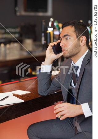 Male executive sipping amber drink talking on smartphone at wooden bar counter notepad, copy space 135362702