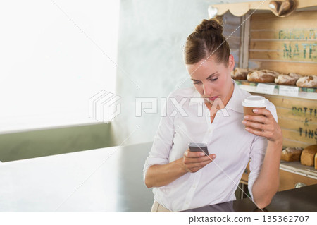 Mid adult woman leaning on bakery counter by window, holding cup, checking smartphone, copy space Mid adult woman leaning on bakery counter by window, holding cup, checking smartphone, copy space 135362707