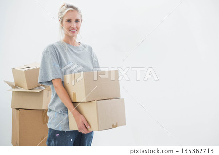 Woman holding two cardboard boxes while wearing paint-splattered T-shirt in white room, copy space 135362713