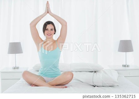 Woman in her 20s sitting on bed in white bedroom practicing yoga with pillows and lamps Woman in her 20s sitting on bed in white bedroom practicing yoga with pillows and lamps 135362715