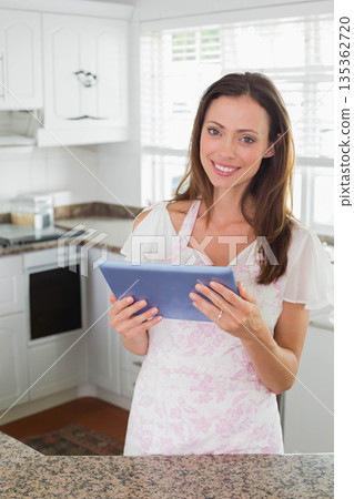 Woman smiling and standing in bright home kitchen holding tablet over granite countertop Woman smiling and standing in bright home kitchen holding tablet over granite countertop 135362720