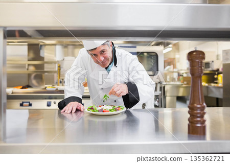 Male chef in uniform garnishing salad with herb sprig on stainless counter with pepper mill, ovens 135362721