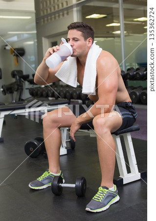 Shirtless man sitting on weight bench drinking from shaker bottle near dumbbells in gym facility Shirtless man sitting on weight bench drinking from shaker bottle near dumbbells in gym facility 135362724