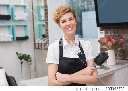 Female hair stylist standing at salon station holding paddle brush with styling tools in apron Female hair stylist standing at salon station holding paddle brush with styling tools in apron 135362731