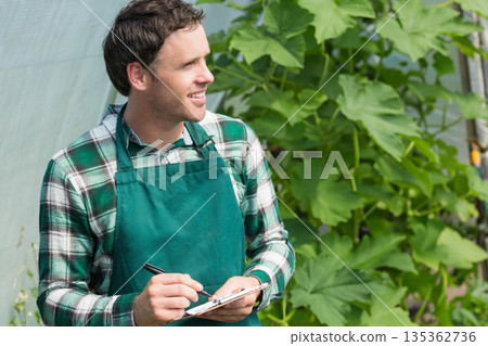Mid-adult man inspecting climbing plants in greenhouse holding clipboard and pen Mid-adult man inspecting climbing plants in greenhouse holding clipboard and pen 135362736