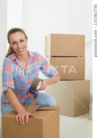 Mid adult woman kneeling on wood floor in entrance area sealing box with handheld tape dispenser 135362738