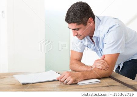 Middle-aged man in shirt leaning on wooden desk in office reviewing papers and notebook, copy space Middle-aged man in shirt leaning on wooden desk in office reviewing papers and notebook, copy space 135362739