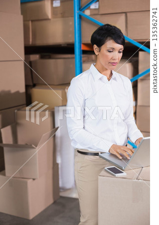 Female warehouse clerk leaning on cardboard boxes checking laptop and smartphone in storage area Female warehouse clerk leaning on cardboard boxes checking laptop and smartphone in storage area 135362741
