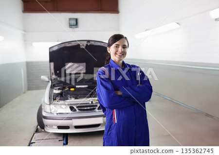 Female mechanic posing in workshop bay in blue coverall near open silver sedan hood showing engine 135362745