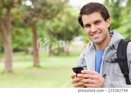 Man standing in leafy park wearing grey hoodie, carrying backpack, checking smartphone, copy space Man standing in leafy park wearing grey hoodie, carrying backpack, checking smartphone, copy space 135362746