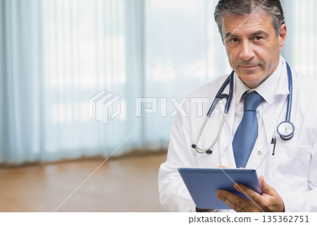 Mature male physician in white lab coat holding tablet in clinic with stethoscope, copy space 135362751