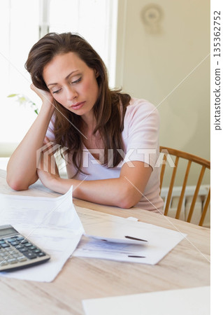 Woman leaning on hand while reviewing documents, pen and calculator at home by wooden dining table Woman leaning on hand while reviewing documents, pen and calculator at home by wooden dining table 135362752