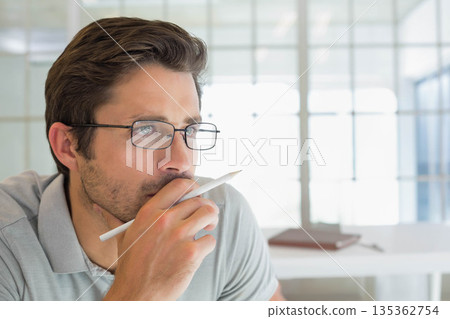 Man wearing eyeglasses and polo shirt holding white stylus near mouth while thinking at office desk Man wearing eyeglasses and polo shirt holding white stylus near mouth while thinking at office desk 135362754