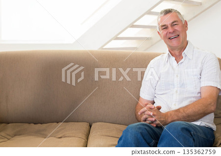 Senior man sitting on beige sofa under skylight with staircase, smiling toward camera, copy space Senior man sitting on beige sofa under skylight with staircase, smiling toward camera, copy space 135362759