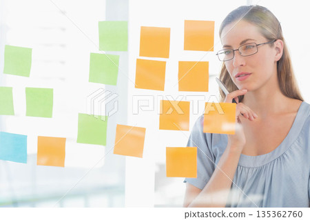Woman wearing eyeglasses holding pen studying sticky notes on glass board in office, copy space Woman wearing eyeglasses holding pen studying sticky notes on glass board in office, copy space 135362760