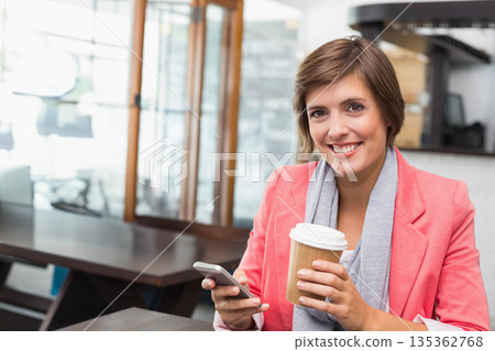 Woman wearing coral blazer holding smartphone and to-go coffee cup at coffee shop table, copy space Woman wearing coral blazer holding smartphone and to-go coffee cup at coffee shop table, copy space 135362768