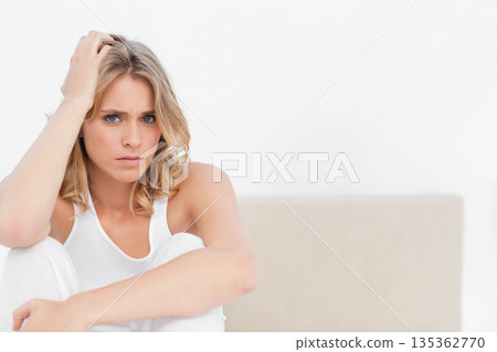 Woman in white tank top gripping hair at scalp, sitting on beige sofa against white wall Woman in white tank top gripping hair at scalp, sitting on beige sofa against white wall 135362770
