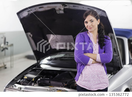 Female standing arms folded beside silver car open hood exposing engine in garage, copy space Female standing arms folded beside silver car open hood exposing engine in garage, copy space 135362772