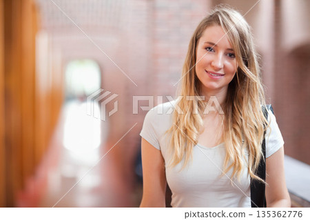 Female student standing in academic corridor with brick wall carrying dark shoulder bag, copy space 135362776