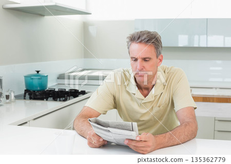 Man sitting and reading newspaper in kitchen at white countertop near turquoise cooking pot and mug Man sitting and reading newspaper in kitchen at white countertop near turquoise cooking pot and mug 135362779