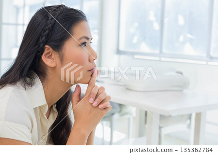 Asian woman sitting at white desk by window, resting chin on fingers beside printer, copy space Asian woman sitting at white desk by window, resting chin on fingers beside printer, copy space 135362784