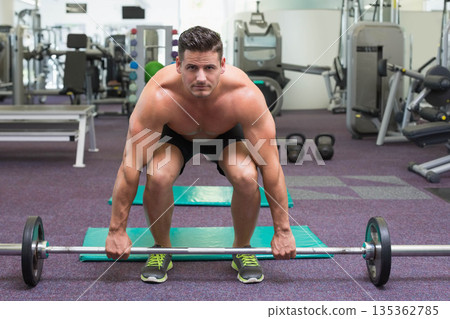 Shirtless man wearing black shorts lifting barbell on mat in gym with kettlebells and mirrored wall Shirtless man wearing black shorts lifting barbell on mat in gym with kettlebells and mirrored wall 135362785