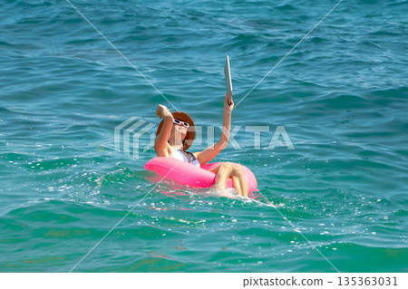 Woman floating water using digital tablet device on bright pink ring during tropical summer vacation holiday. Woman floating water using digital tablet device on bright pink ring during tropical summer vacation holiday. 135363031