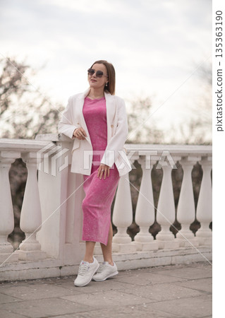 Woman PinkDress Balcony: Woman poses outdoors on a balcony, wearing sunglasses in pink dress and white jacket. Woman PinkDress Balcony: Woman poses outdoors on a balcony, wearing sunglasses in pink dress and white jacket. 135363190