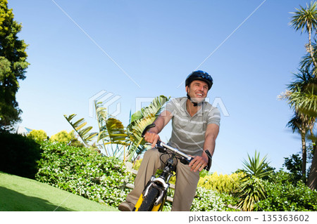 Mid-adult man riding bicycle wearing striped polo shirt and helmet along tropical garden pathway 135363602