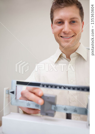 Male health technician wearing light shirt standing behind beam scale adjusting weight in clinic 135363604