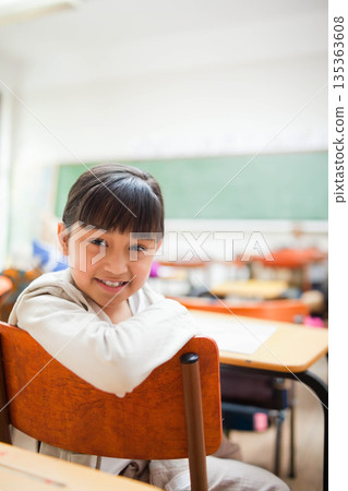 Schoolgirl sitting backward on chair by orange-topped desks near chalkboard in classroom Schoolgirl sitting backward on chair by orange-topped desks near chalkboard in classroom 135363608