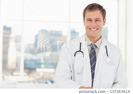 Male doctor smiling while standing in clinic by window with stethoscope and lab coat, copy space 135363614