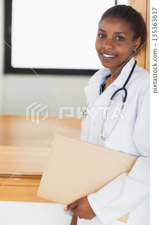 Female doctor holding file folder and stethoscope leaning in clinic near window shade, copy space Female doctor holding file folder and stethoscope leaning in clinic near window shade, copy space 135363617