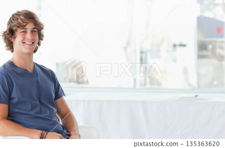 Man wearing V-neck shirt sitting on chair in lounge by table and large windows, copy space 135363620