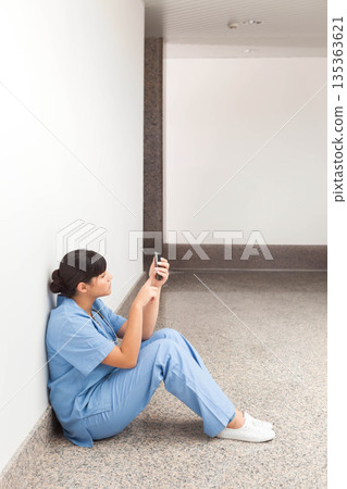 Asian female nurse in scrubs sitting on hospital corridor floor checking smartphone, copy space 135363621