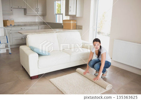 Woman in her twenties crouching in living room rolling up beige rug by window, copy space Woman in her twenties crouching in living room rolling up beige rug by window, copy space 135363625