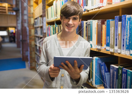 Man standing in narrow library aisle holding tablet and browsing bookshelves under soft lighting 135363626