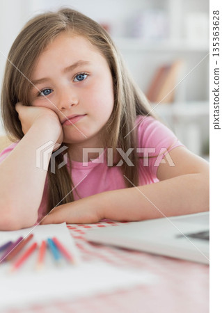 School-age girl resting cheek on hand at home study desk with colored pencils, paper and laptop School-age girl resting cheek on hand at home study desk with colored pencils, paper and laptop 135363628