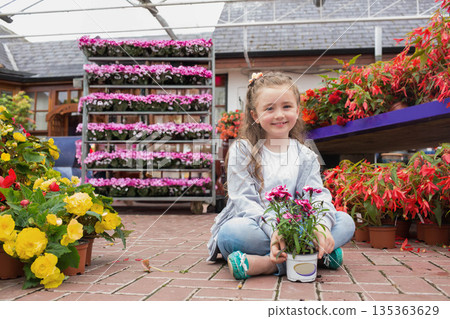 Female child sitting cross-legged holding white pot with pink blossoms on brick floor in greenhouse Female child sitting cross-legged holding white pot with pink blossoms on brick floor in greenhouse 135363629