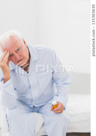 Senior man wearing pajamas sitting on bed holding prescription pill bottle and pressing forehead Senior man wearing pajamas sitting on bed holding prescription pill bottle and pressing forehead 135363630