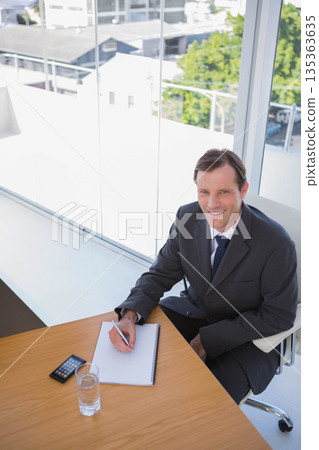 Man in business suit writing in notebook at office desk with smartphone, water glass, copy space 135363635