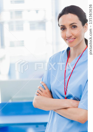 Female nurse standing arms crossed in clinic wearing blue scrubs with red lanyard, copy space 135363636
