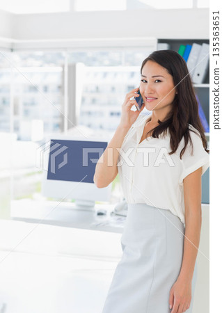 Asian woman standing by office desk using smartphone near computer and binders, copy space Asian woman standing by office desk using smartphone near computer and binders, copy space 135363651