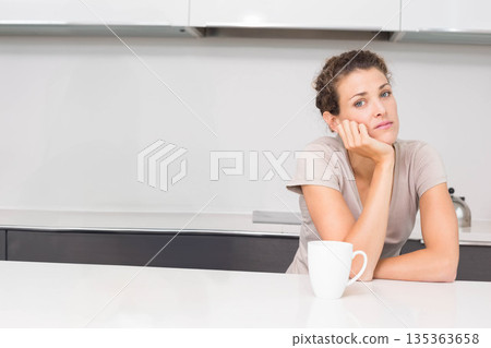 Curly-haired woman resting chin on hand at kitchen countertop with mug and kettle, copy space Curly-haired woman resting chin on hand at kitchen countertop with mug and kettle, copy space 135363658