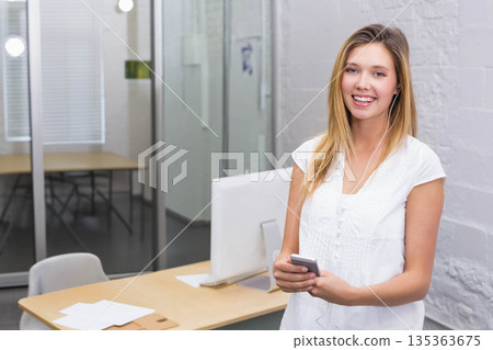 Female office employee standing by desk with monitor holding smartphone and earphones copy space Female office employee standing by desk with monitor holding smartphone and earphones copy space 135363675
