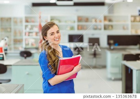 Smiling female student wearing blue scrubs and holding red spiral notebook in science lab classroom 135363678