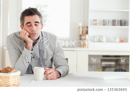 Man in early 30s resting head on hand, sipping from white mug at bright kitchen table Man in early 30s resting head on hand, sipping from white mug at bright kitchen table 135363693